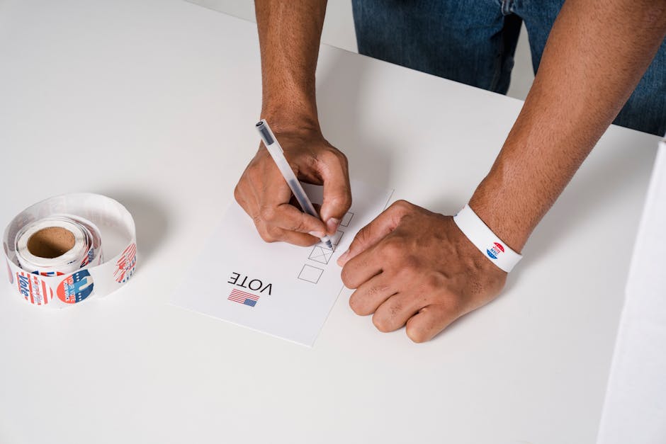 A person wearing a wristband fills out a voting ballot with a pen. Voting stickers visible.
