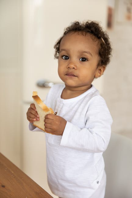 A cute child holding a slice of bread, standing indoors, looking curious.