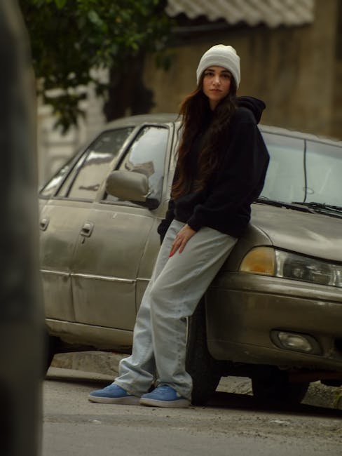 Casual portrait of a young woman leaning on a car, wearing casual clothing and a beanie, in an urban environment.