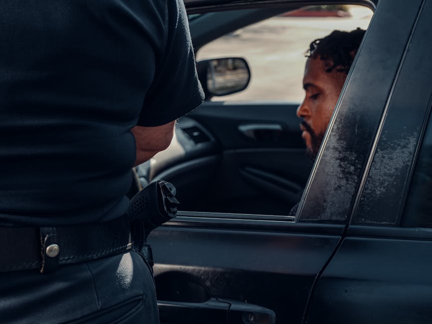 A police officer issuing a ticket to a man seated in a car, captured outdoors in daylight.