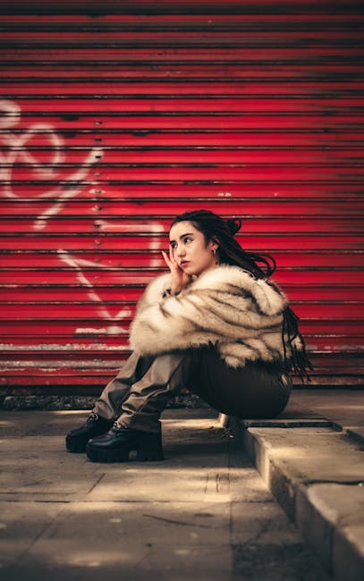 Young woman in stylish attire sits against a red urban background, exuding confidence and style.