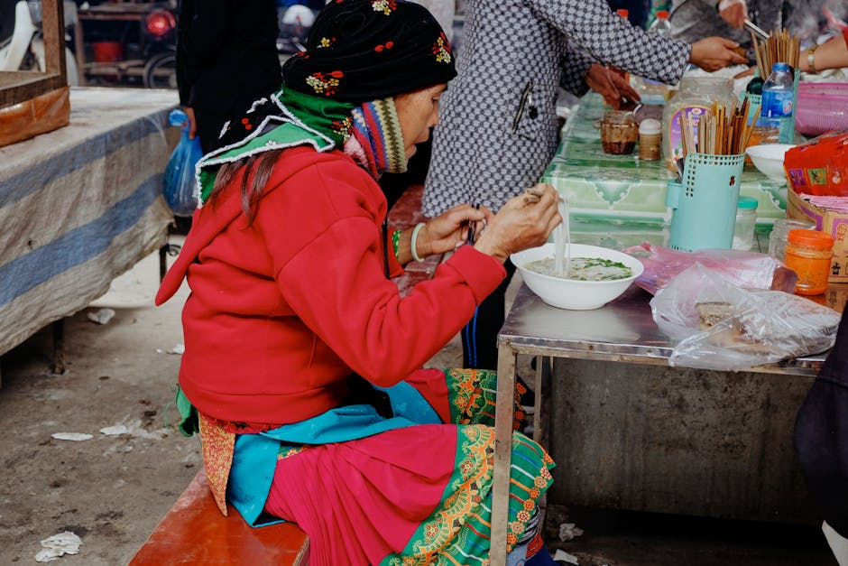 An elderly woman in traditional clothing enjoys noodles at an outdoor market table.