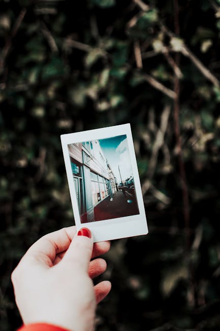A hand with manicured nails holds an instant photo against a green outdoor background.