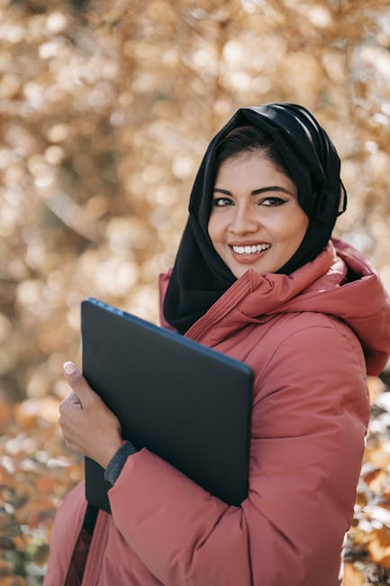 Cheerful Muslim woman with a laptop in an autumn park, symbolizing remote work and positivity.