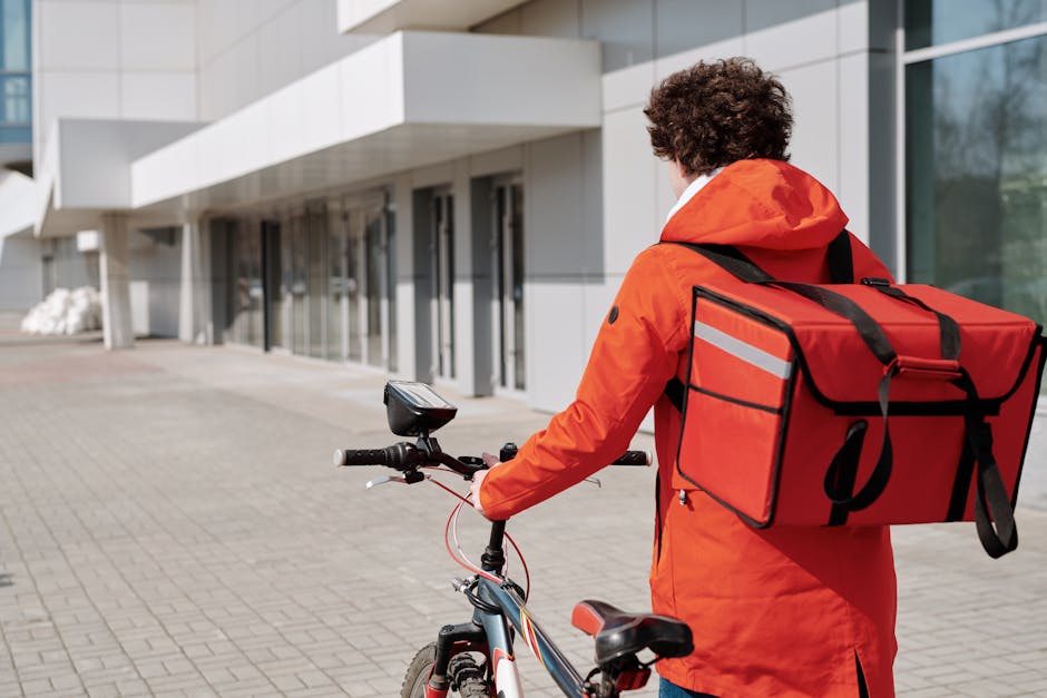 A bicycle courier in an orange jacket and delivery bag on a city street, ready for delivery.