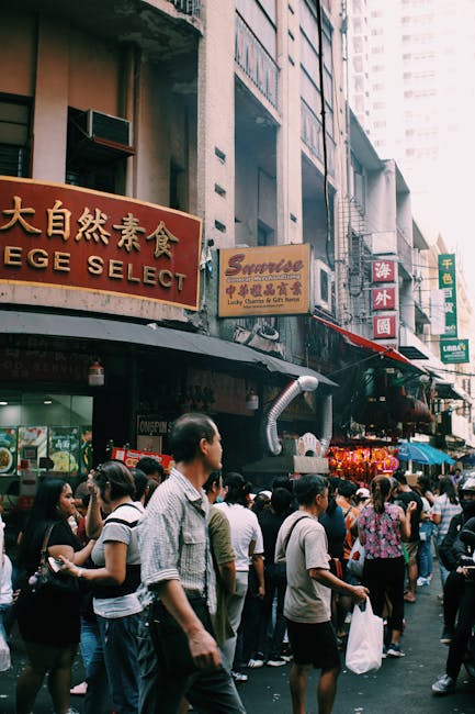 Busy urban street market scene with diverse crowd and signs in various languages.