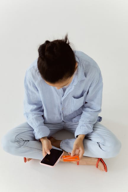 Woman in blue attire holding smartphone seated on white surface with card.