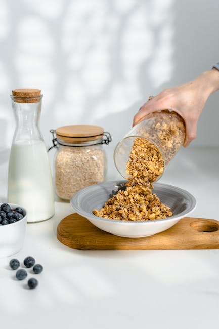 Close-up of granola being poured into a bowl with milk and blueberries for a healthy breakfast setup.
