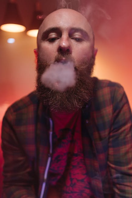 A bald man with a beard exhales smoke in a cozy indoor setting with ambient lighting.