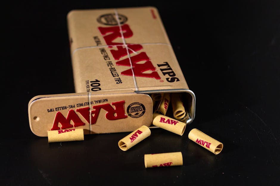 Close-up of RAW pre-rolled tips spilling from a metal tin on a black surface.
