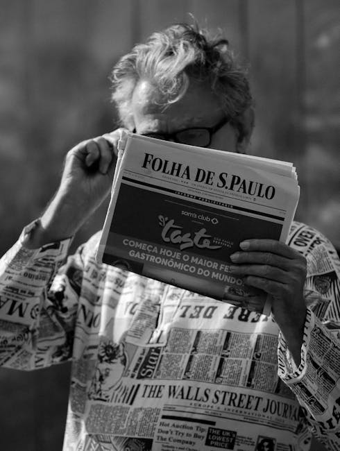 Black and white photo of a man reading Folha de S.Paulo newspaper outdoors, wearing a newsprint shirt.