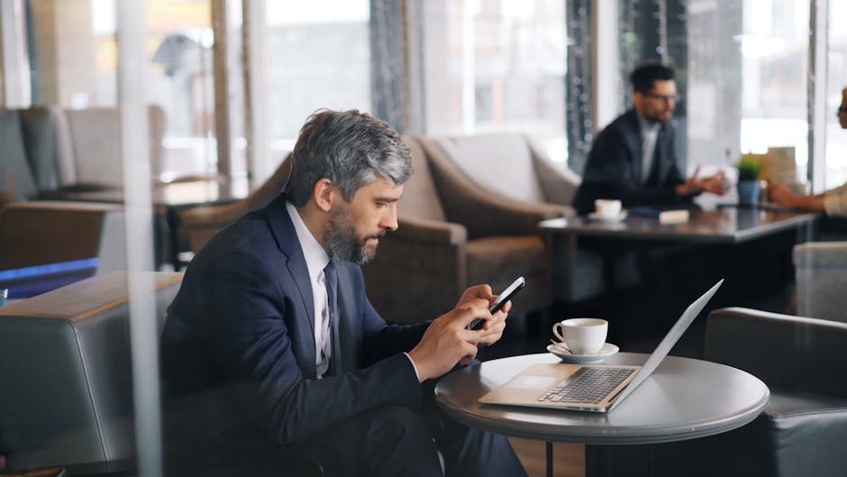 Professional man using smartphone and laptop in a café setting.