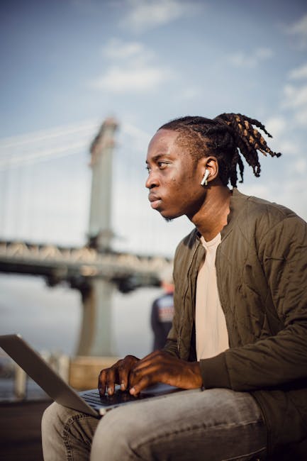 A focused young man working on his laptop outdoors with earphones near a bridge.
