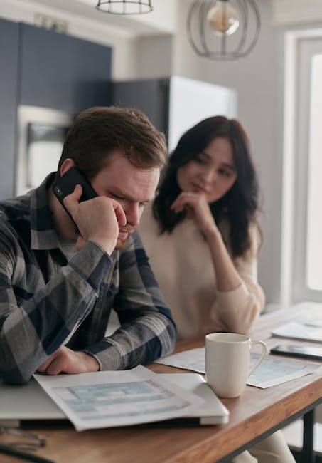 A worried couple sits at a table with bills and a phone, planning their finances.