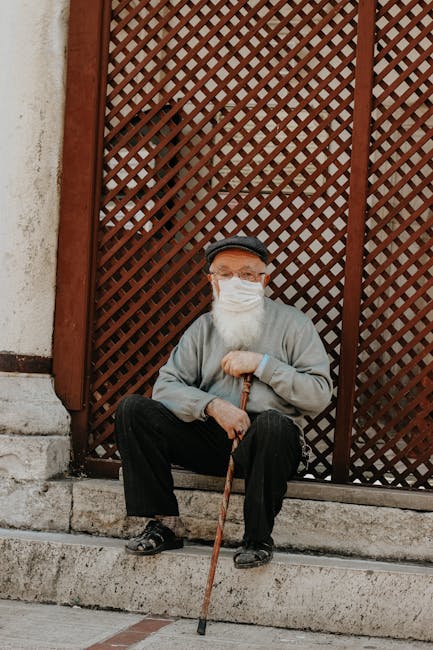 Senior man with face mask seated outdoors against a lattice fence, grasping a walking stick.