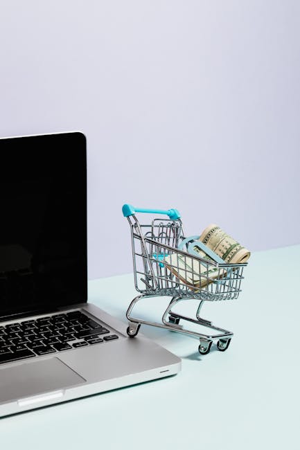 Mini shopping cart with rolled cash next to a laptop, representing online shopping.