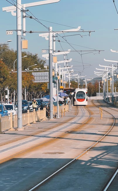 A modern tram approaches a city station surrounded by trees and vehicles, under a clear blue sky.