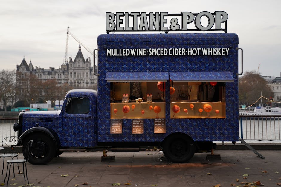 Colorful food truck serving mulled wine, spiced cider, and hot whiskey by the riverside with cityscape backdrop.