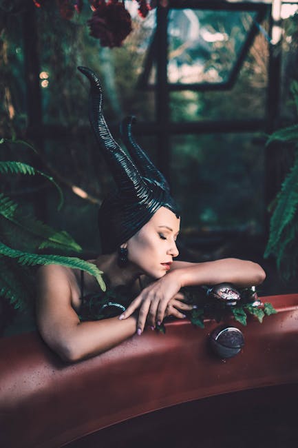 Woman in dark fantasy costume with horns relaxing by a bathtub amidst lush greenery.