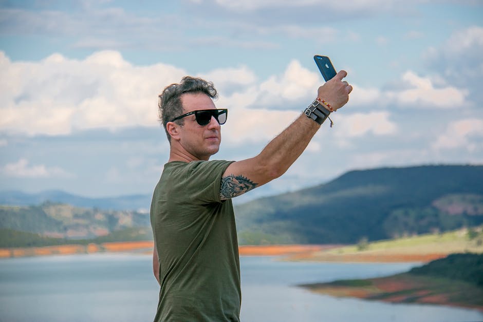 Man taking a selfie outdoors by the lake with scenic background, capturing the essence of travel and adventure.