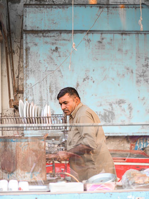A man washing dishes at an outdoor street vendor stall, amidst vibrant colors and rustic surroundings.