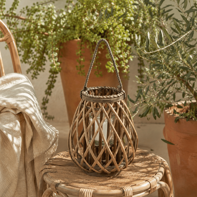 A small bulbous wicker lantern styled on a low rattan side table on a sun-drenched terrace, surrounded by terracotta pots with trailing greenery and a linen throw draped over a nearby chair. Soft afternoon light, relaxed and organic mood.