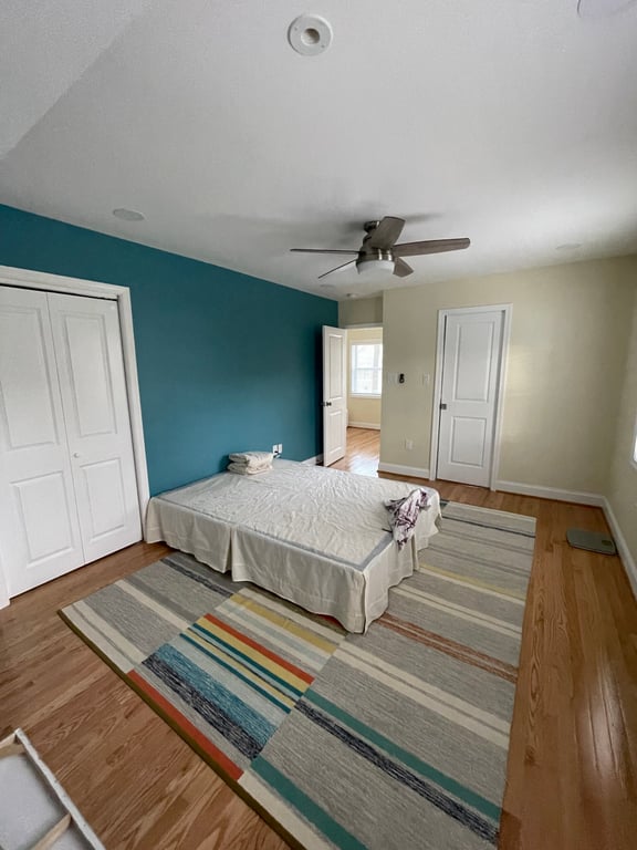 Minimally furnished bedroom with hardwood floors, striped rug, teal accent wall, ceiling fan, and closets