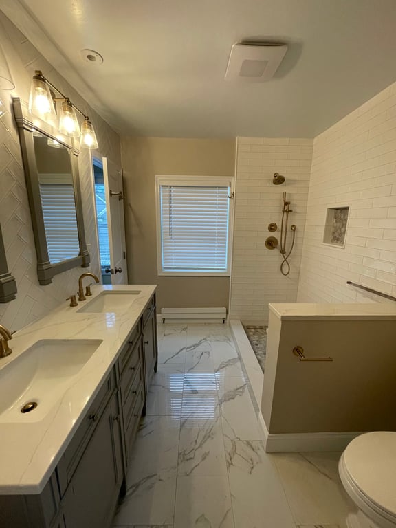 Modern bathroom with marble floors, double-sink vanity with gold fixtures, large mirrors, walk-in shower with shelf, and tiled walls