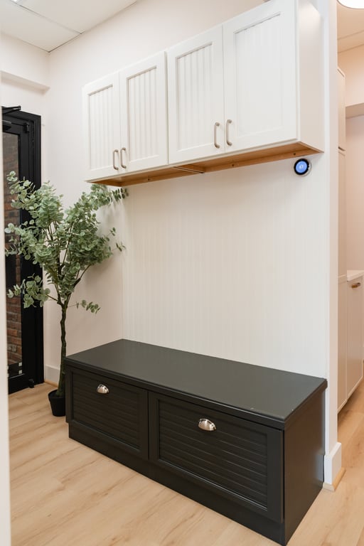 Modern mudroom with dark storage bench, white upper cabinets, wooden shelf, wood flooring, and a potted faux tree