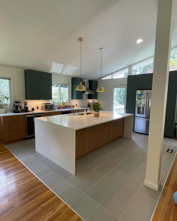 Modern kitchen with large white island countertop, wood and dark green cabinets, stainless steel appliances, and gold pendant lights
