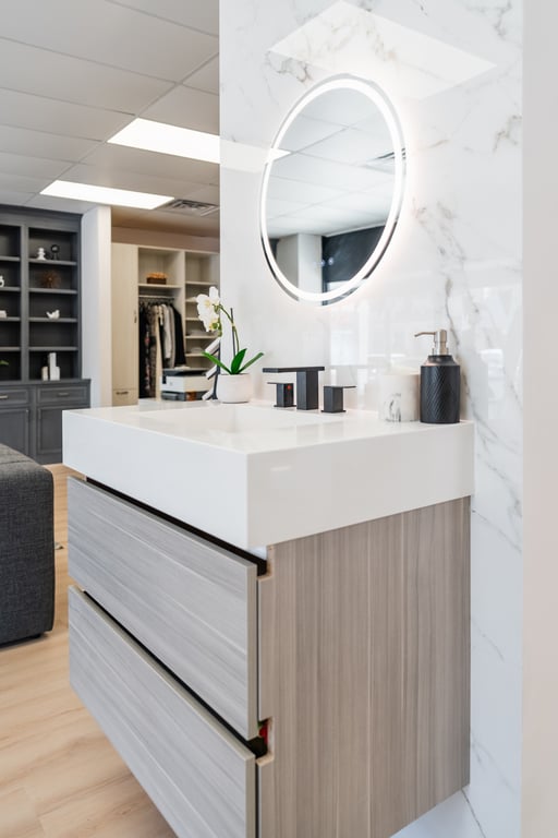 Modern bathroom vanity with light wood drawers, white countertop, and round backlit mirror against a marble wall in the Bethesda showroom