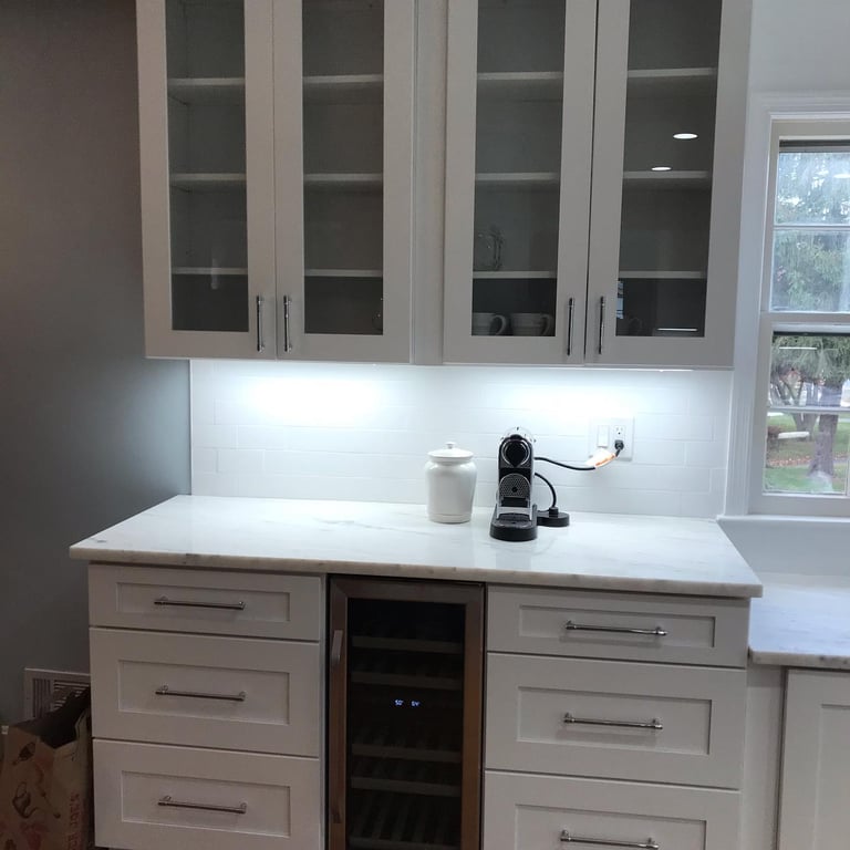 Modern kitchen countertop with white cabinets, small glass-front wine fridge, and tiled backsplash with greenery view