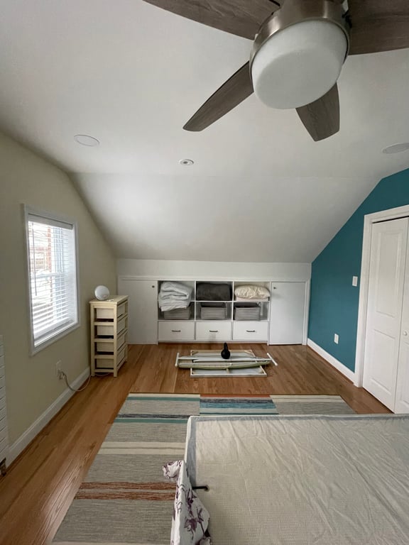 Minimalist bedroom with slanted ceiling, hardwood floor, teal accent wall, ceiling fan, low cube storage, and natural light