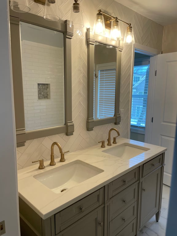 Modern bathroom with double sink vanity, gold faucets, rectangular mirrors, wall-mounted lights, and herringbone tile backsplash