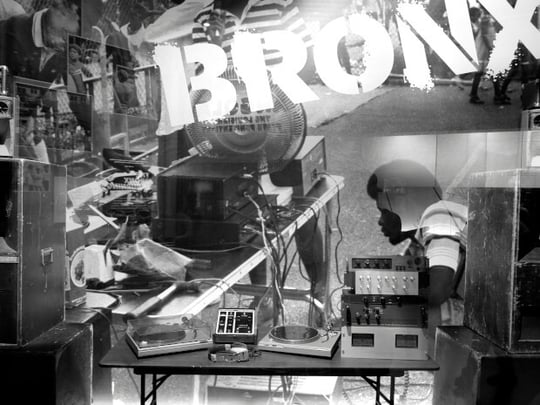 Black and white image of a person sat behind speakers and audio equipment