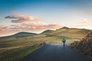 A tarmac road with green hills in the background and a cyclist in the foreground. A few clouds are in a blue sky.