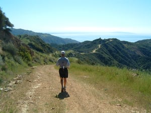 A stock image of a trial runner on a gravel trail with mountains in the background and grass to the side of the path