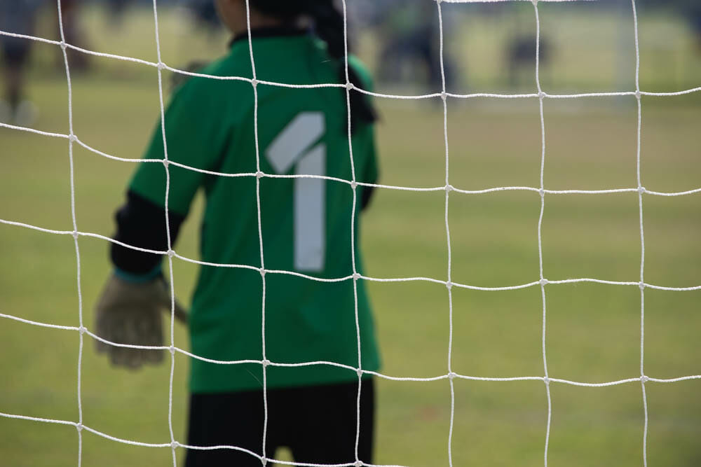 Close up football goal net, seeing a young girl soccer goalkeeper watching her teammate playing. Youth Soccer game on a sunny summer school tournament Day. A football match going on in a background.
