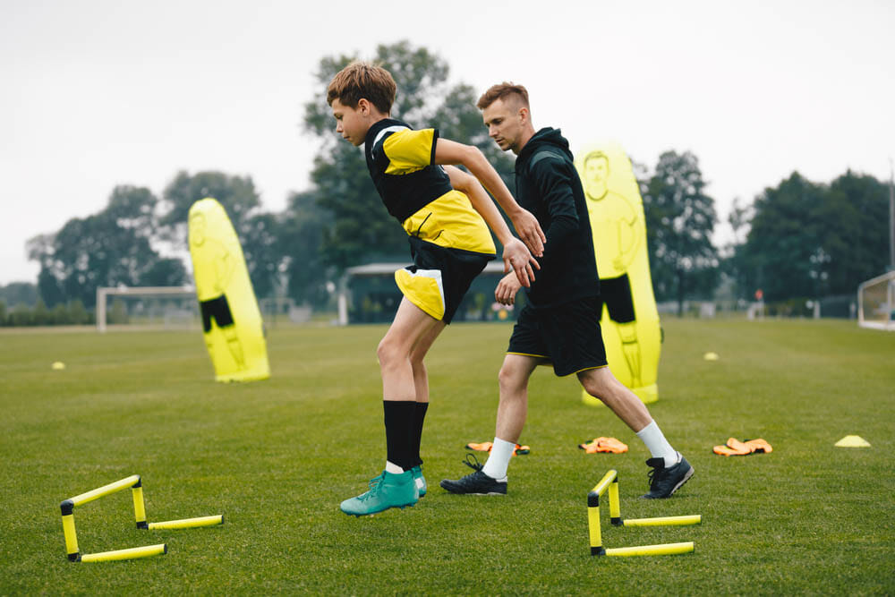 Young soccer athlete practicing strength and agility skills at soccer school academy. Soccer coach coaching a young player. Teenage boy jumping over hurdles. Boy with personal trainer.