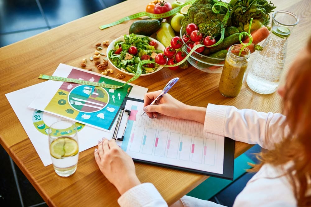 Woman dietitian in medical uniform with tape measure working on a diet plan sitting with different healthy food ingredients in the green office on background. Weight loss and right nutrition concept
