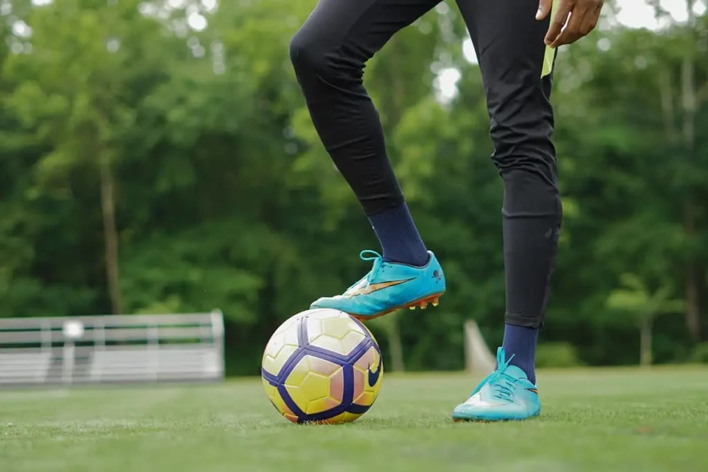 A pair of legs with one stood on a football. Wearing blue football boots on grass