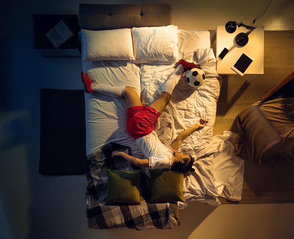 Kicking. Top view of young professional football, soccer player sleeping at his bedroom in sportwear with ball. Loving his sport, workaholic, playing match even if resting. Action, motion, humor.