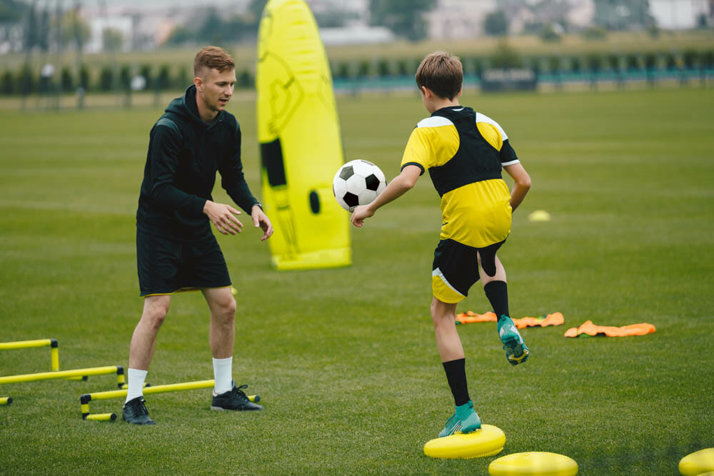 Young Man Coaching Soccer Boy. Coach on Training Session With Teenage Boy. Soccer Player Kicking Ball Standing on Stability Cushion on Grass Field. Football Player Improving Skills With Trainer