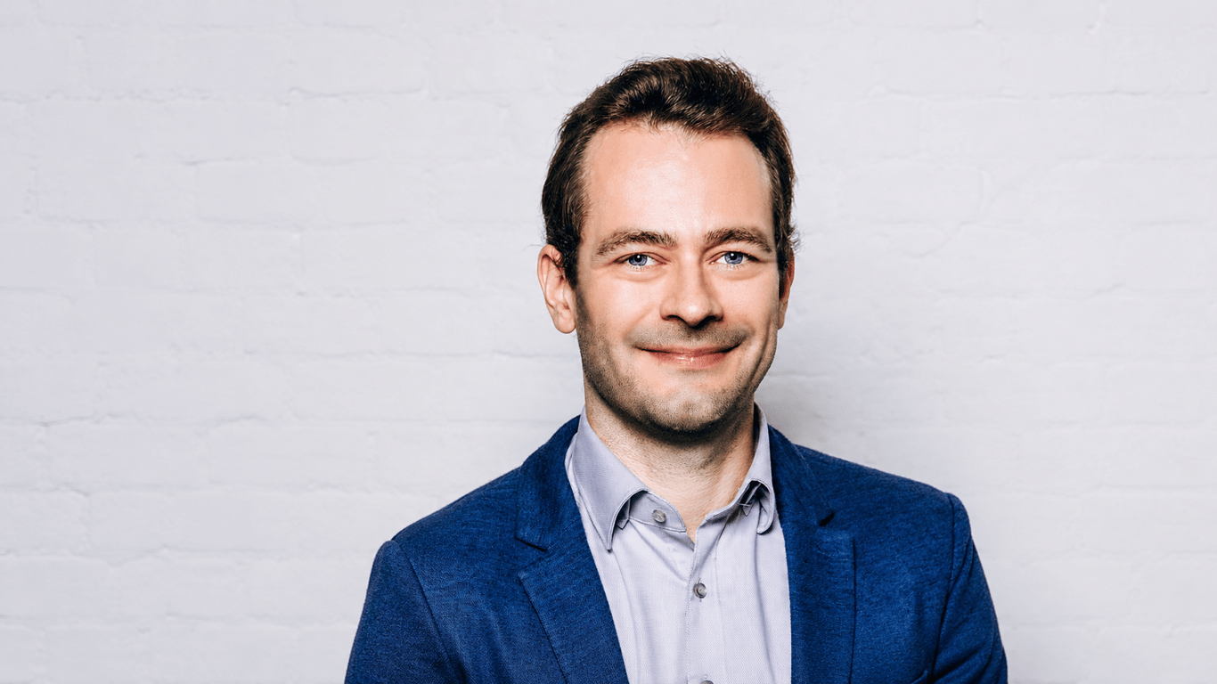 Professional 16:9 portrait of Former EF researcher Dankrad Feist in a blue blazer and light shirt, smiling against a clean white brick background.