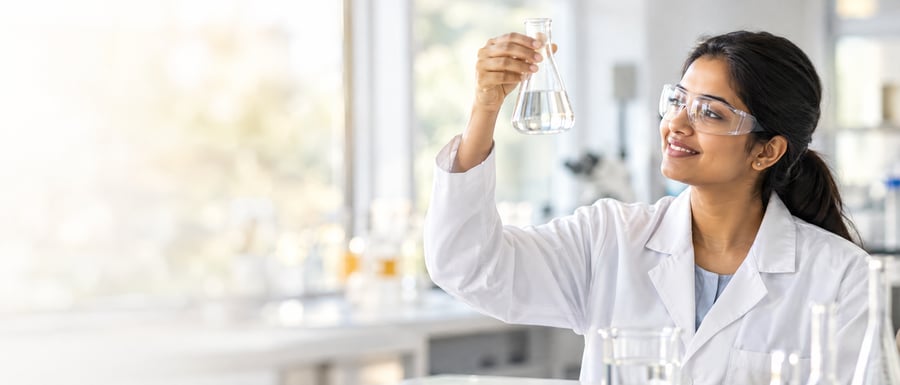 A scientist in a bright laboratory holds a flask of clear liquid up to the light