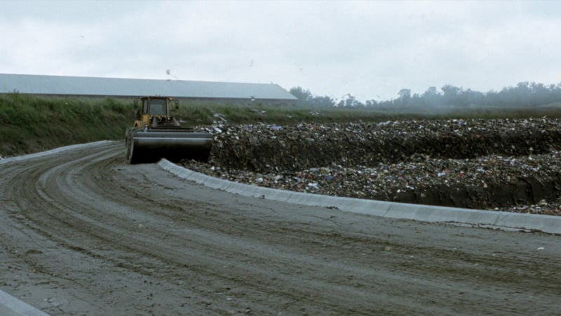 A tracked compactor working a terraced cell of covered waste at a landfill, a muddy haul track leading up to it