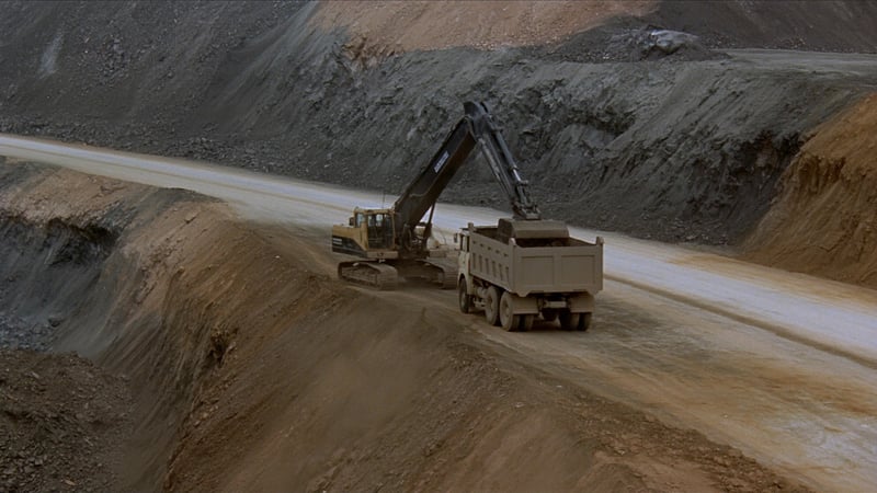 An excavator loading a rigid dump truck on a freshly cut mine bench, a temporary haul road curving across raw earth below a benched highwall