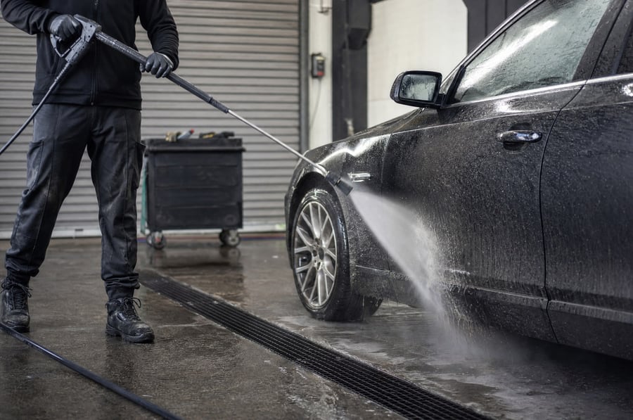 A worker pre-washing a dirty car with a high pressure lance in a vehicle workshop, a fan jet of water lifting road dirt off the front wheel arch, wet concrete floor with a drain