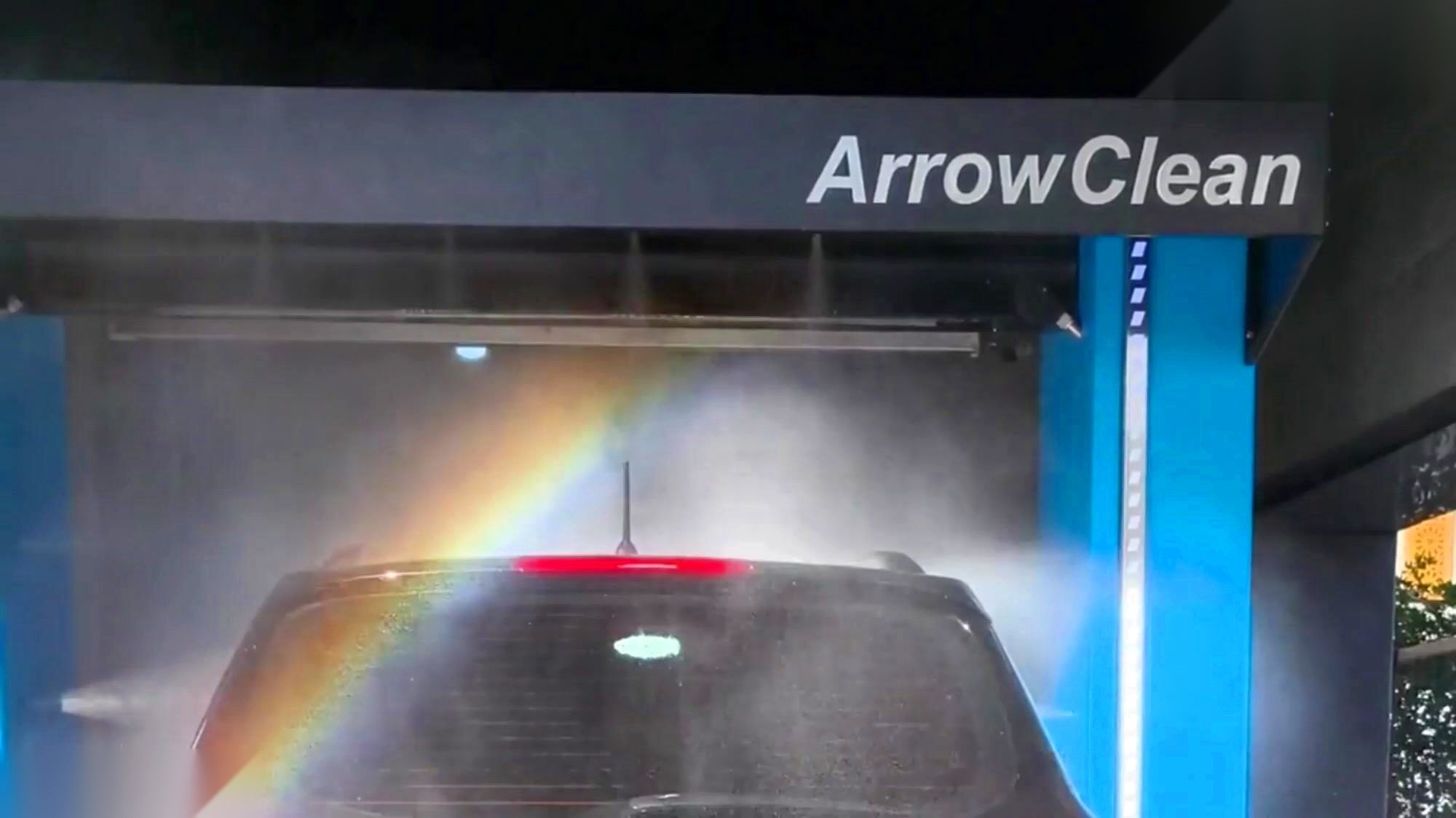 The KKE ArrowClean gantry over a car mid-wash, its name along the beam, spray falling from the top bar and a rainbow through the mist