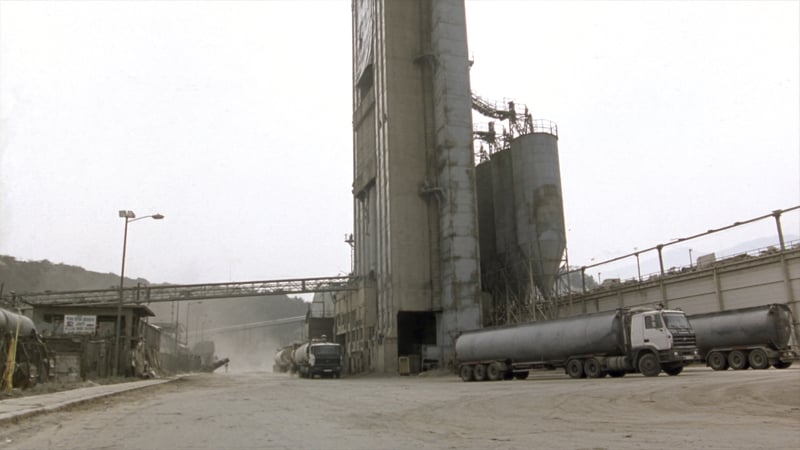 A cement works yard under limestone dust, preheater tower and clinker silos above bulk tankers